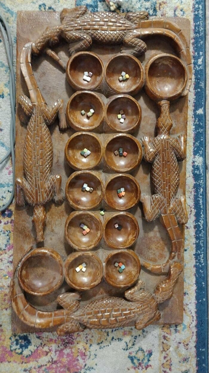 A carved wooden board game, possibly Mancala, featuring crocodile motifs. Small colorful beads fill the circular depressions. Unusual possessions.