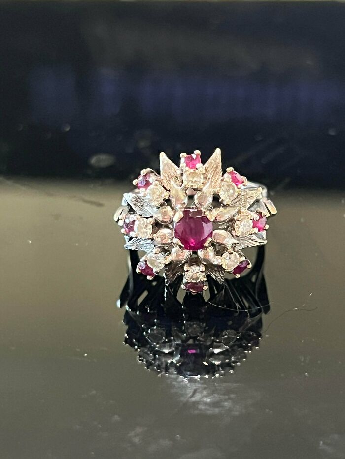 A silver ring with a cluster of red and white gemstones reflecting on a dark surface, highlighting its unusual possessions.