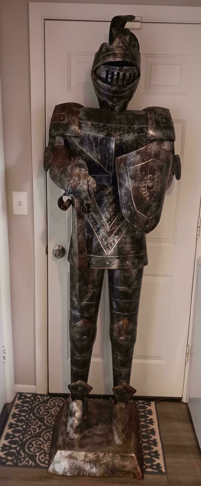 An aged, full-body knight's armor with a shield and sword, an unusual possession, standing by a white door.