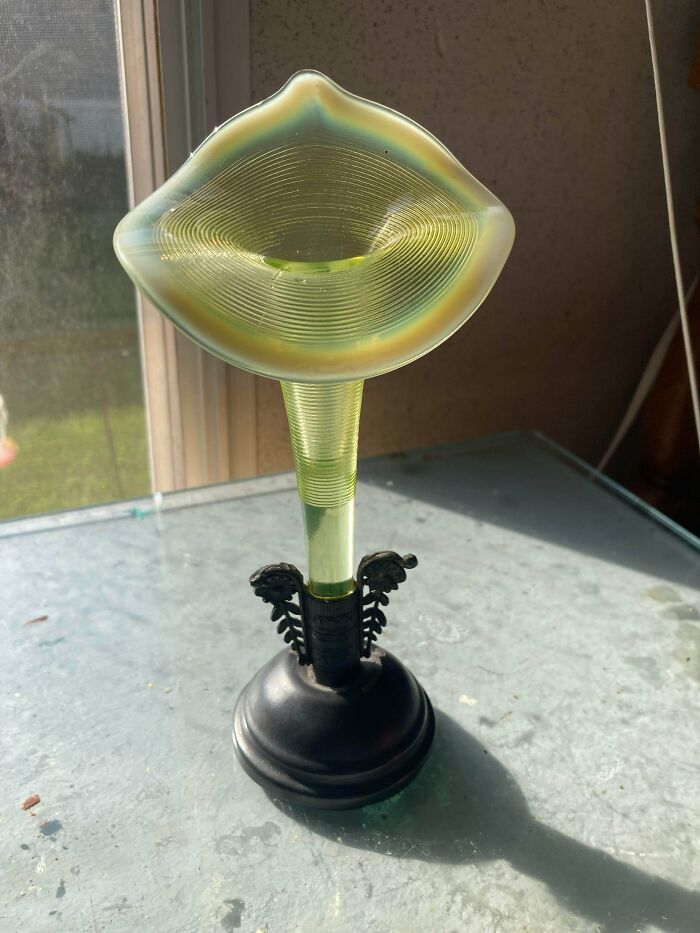 An iridescent green glass vase with a wavy rim and ribbed interior, held by a black ornate base, an unusual possession.