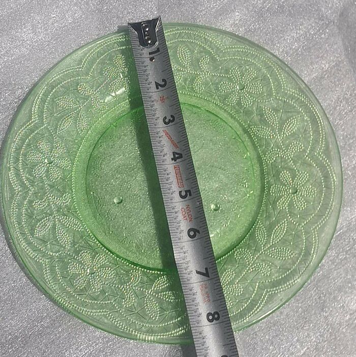 A measuring tape across a light green depression glass plate with an intricate floral pattern, highlighting its unusual possessions value.