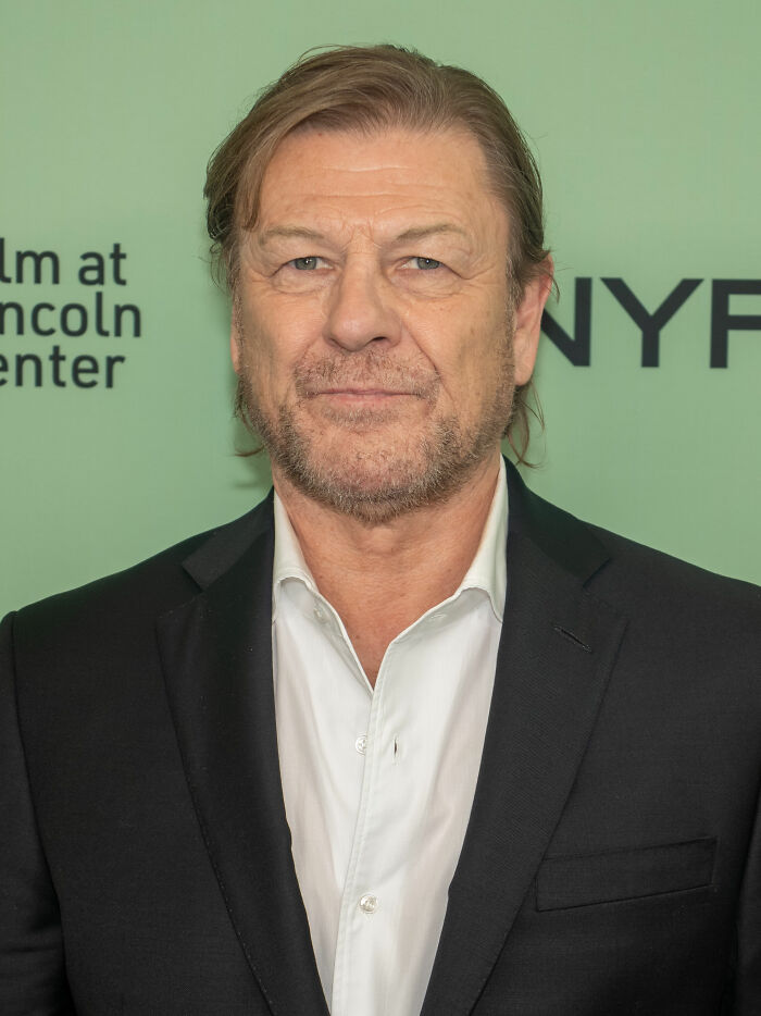 Sean Bean wearing a black suit and white shirt, posing at an event with a green background.