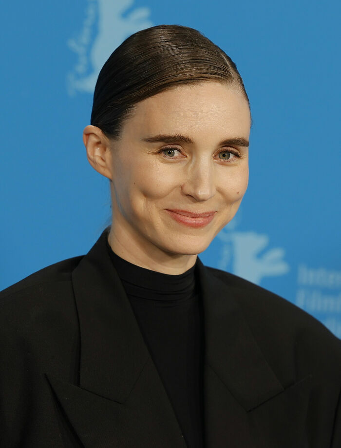 Rooney Mara smiling at a film festival event, wearing a black outfit with sleek hair against a blue backdrop.