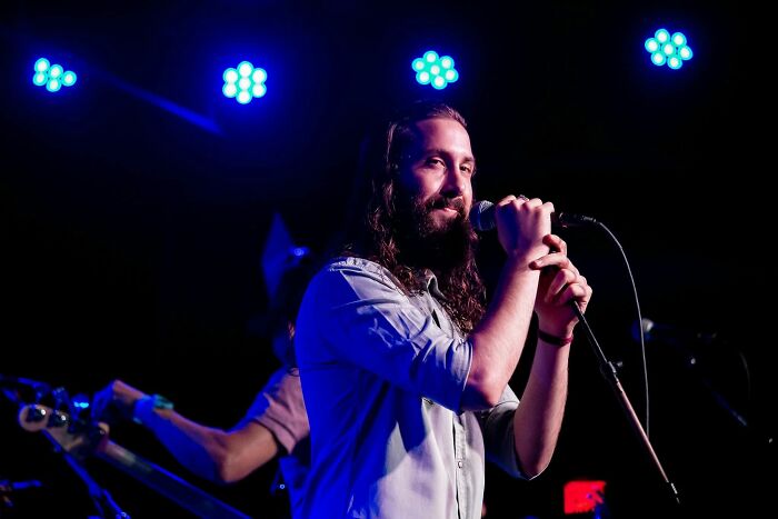 Avi Kaplan performing on stage, holding a microphone, with blue stage lights in the background.