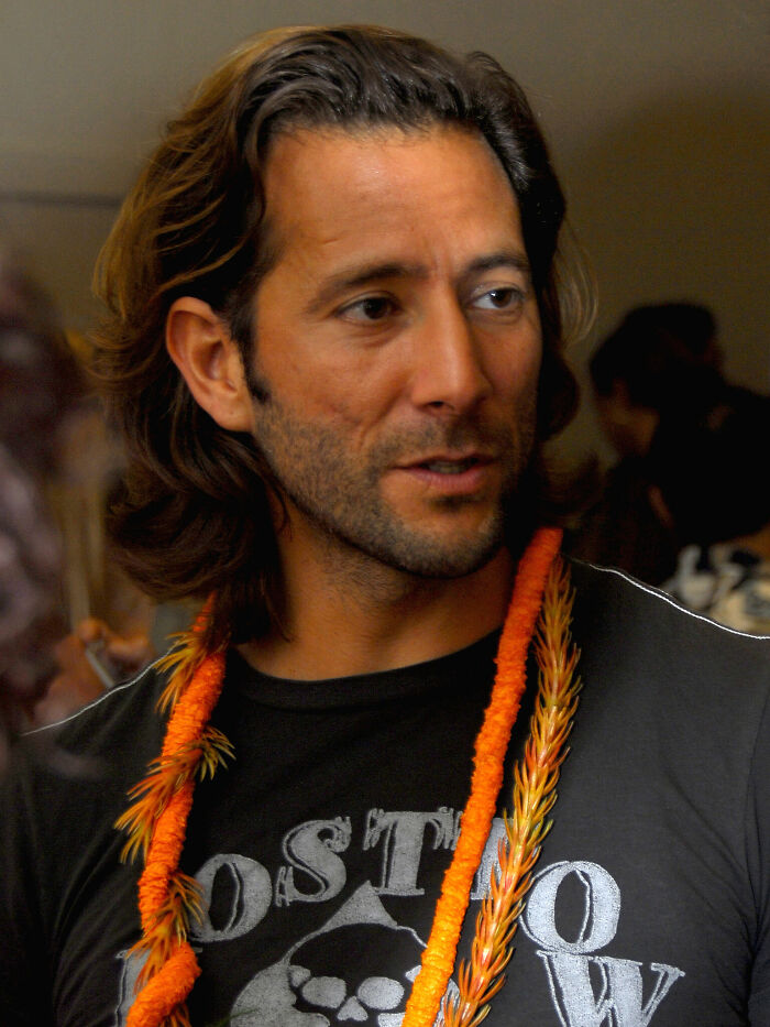 Henry Ian Cusick wearing a black graphic tee and orange lei, captured in a candid moment at an indoor event.