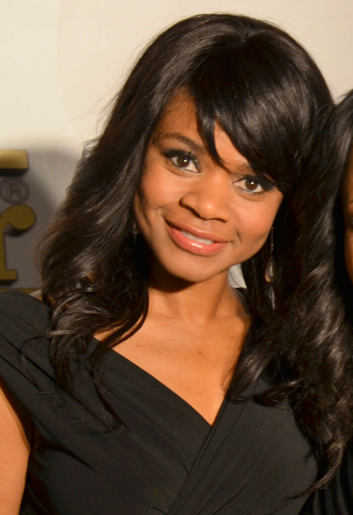 Kimberly Elise smiling, wearing a black dress and earrings, with styled hair in a warm indoor setting.