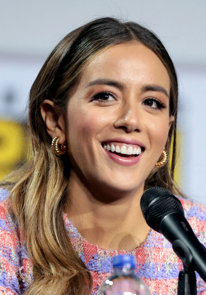Chloe Bennet smiling and speaking into a microphone at an event, wearing gold hoop earrings and a colorful top.