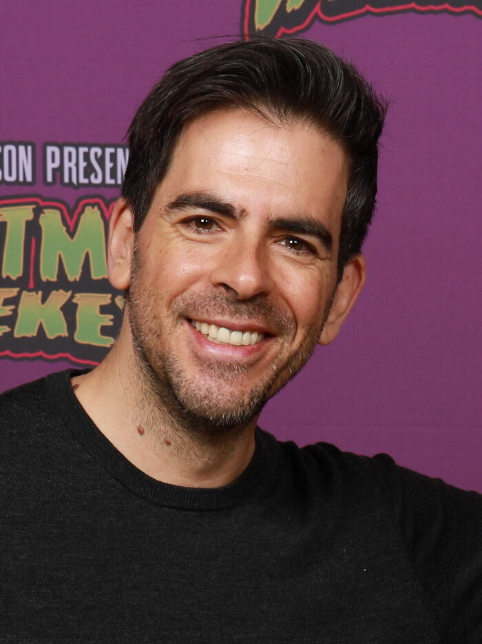 Eli Roth smiling at an event, wearing a black sweater with a colorful background, highlighting his bio and career.