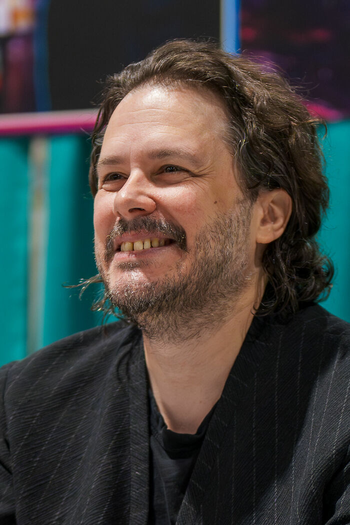 Edgar Wright smiling with casual hair and beard, wearing a dark jacket in an indoor setting.
