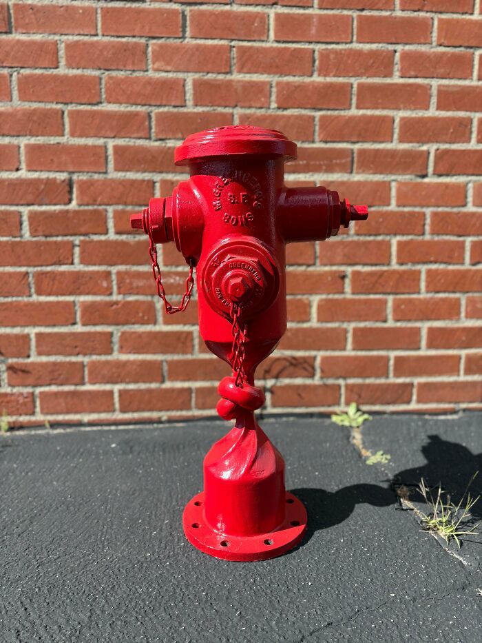 A red fire hydrant twisted at its base against a brick wall, showcasing a handmade project with talent and magic.