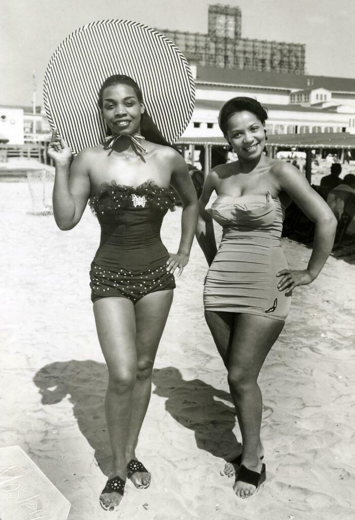 Two women in vintage swimsuits posing on a sunny beach, reflecting decades-old fashion in classic black and white photography.