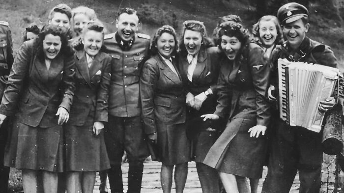 Group of smiling people in vintage military attire, celebrating with an accordion in a historical photo representing a vastly different world.