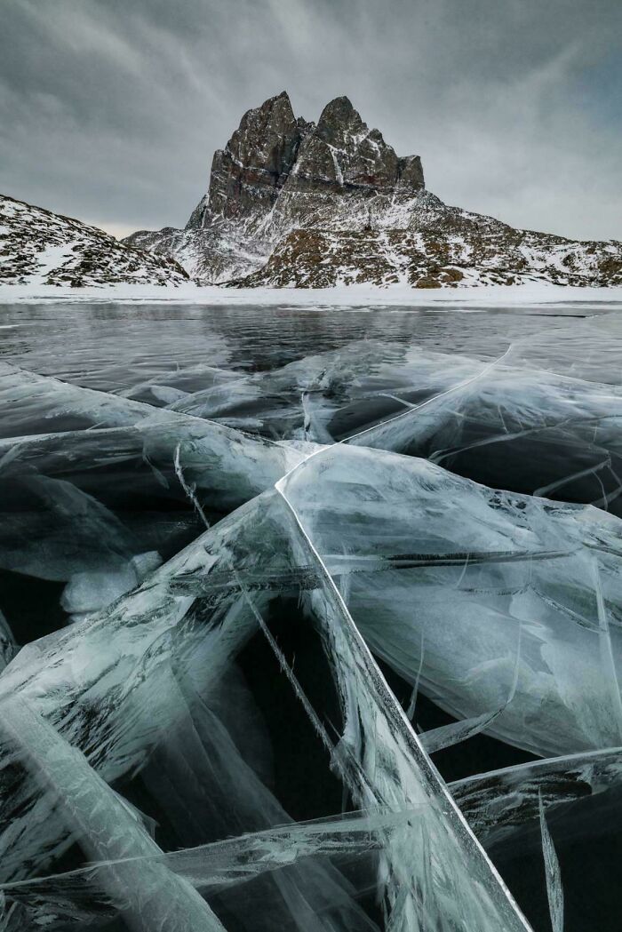 Sharp, geometric ice cracks on a frozen lake with a majestic, snow-dusted mountain under a cloudy sky. Satisfying pictures for a reset.