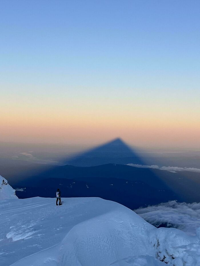 A person standing on a snowy mountain peak at sunrise, casting a huge triangular shadow across the valleys. Satisfying picture for a gentle reset.