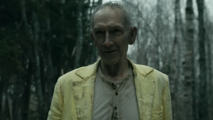 Homem mais velho com uma jaqueta amarela parado em uma floresta escura, sugerindo uma reviravolta aterrorizante na estreia da 4ª temporada.