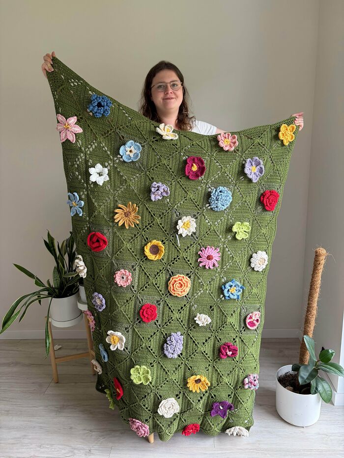A woman holding up a large green crochet blanket adorned with colorful handmade flowers, showcasing handmade projects.
