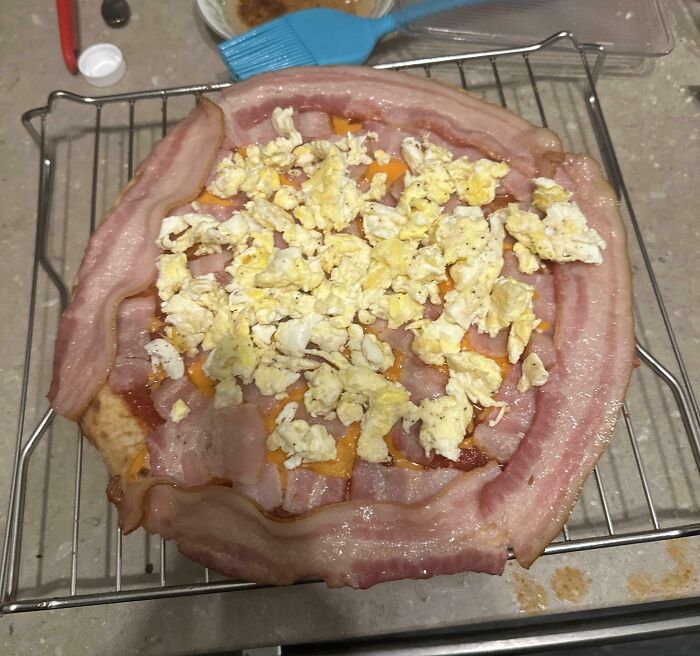 An unbaked Bananasausage pizza topped with raw bacon crust, scrambled eggs, and cheese on a wire rack.