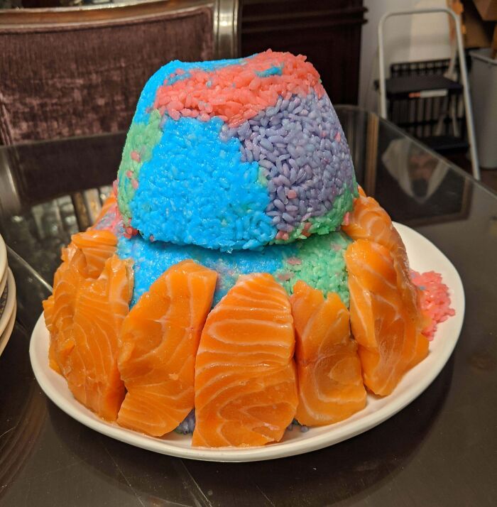 A vibrant rice cake, shaped like a volcano, with blue, pink, and green rice, surrounded by raw salmon slices. A bananasausage food crime.