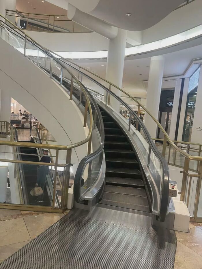 Mall interior with an empty escalator surrounded by glass railings and white architectural columns.