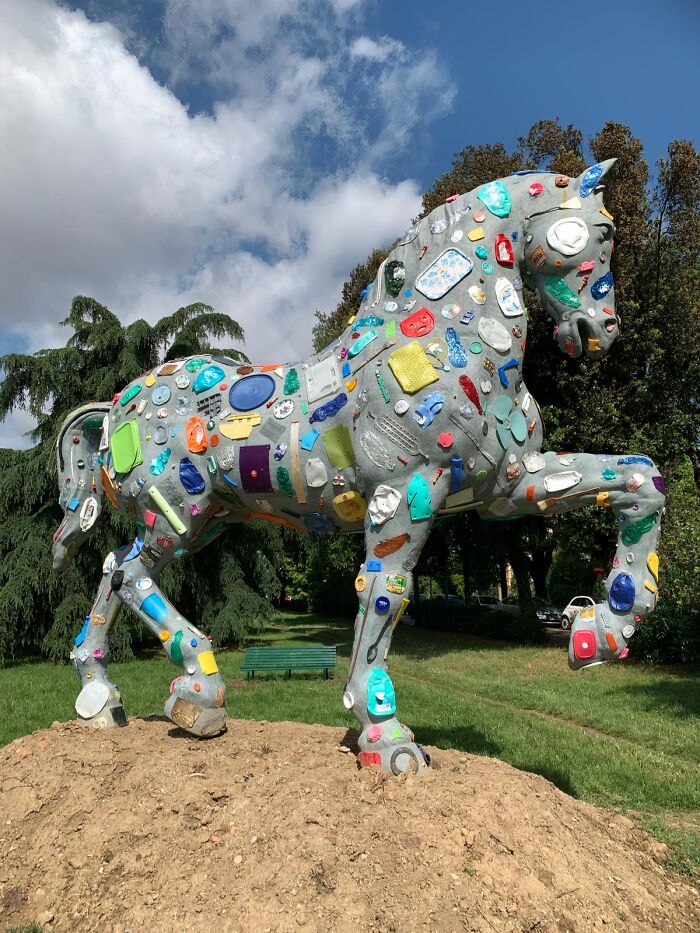 A large horse sculpture made from recycled trash and various useful or artistic items, standing on a mound in a park.
