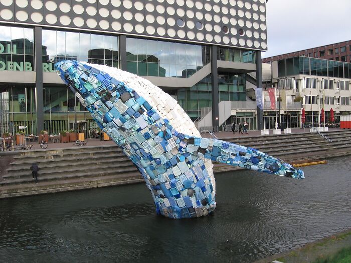 A large whale sculpture made from recycled blue and white trash rising from water, a creative way to use trash.