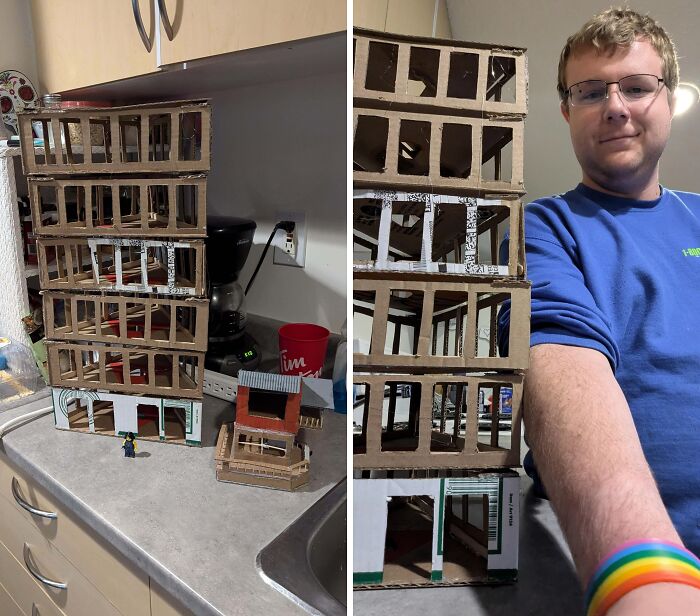 A man proudly stands beside a tall structure made from cardboard, showcasing a creative trash art project.