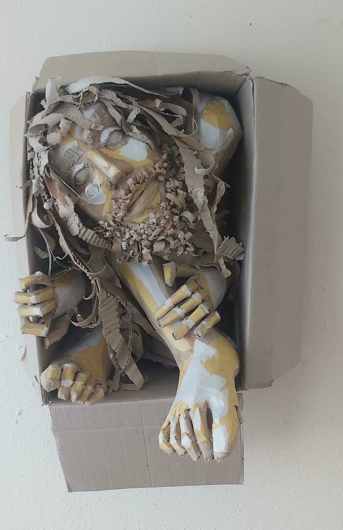 A sculpture of a figure made from cardboard and paper, painted yellow and white, emerging from a box. Trash art.