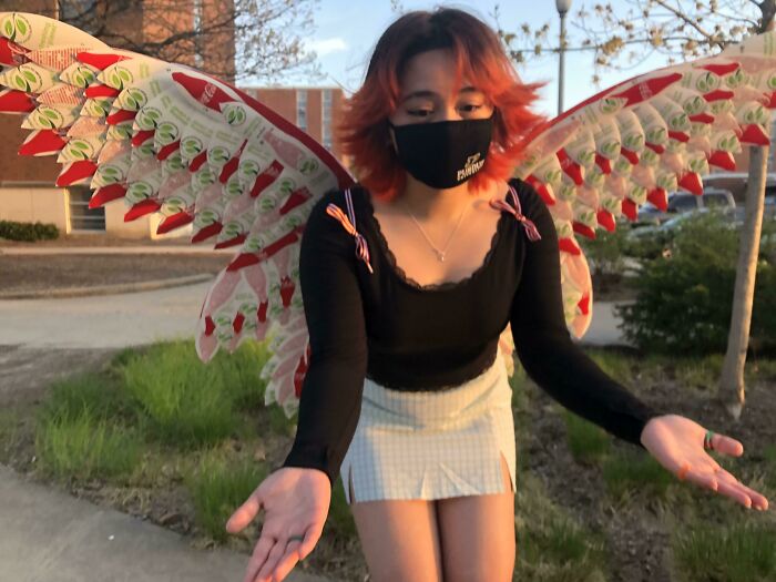 A young woman in a black mask and shirt, with orange hair, wearing creative people wings made from trash.