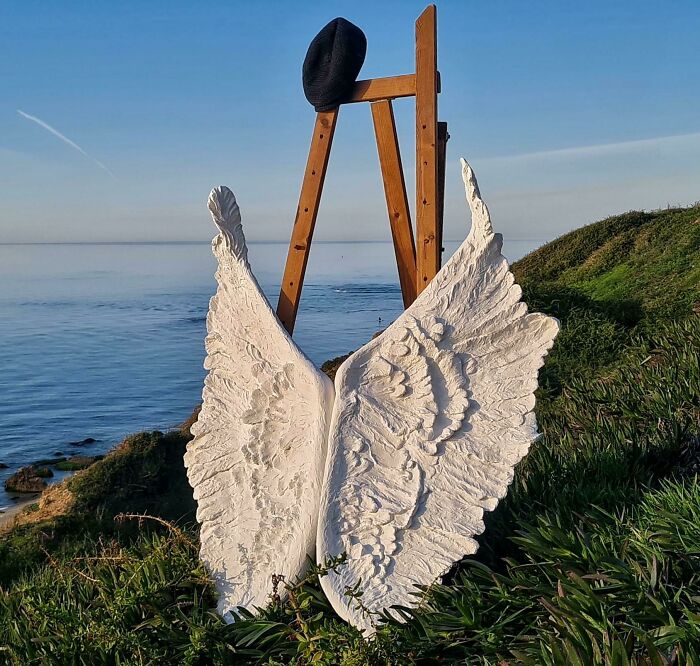 White angel wings made from trash, set on a grassy cliff with an easel and beret overlooking the ocean. Creative people turned trash into something useful.