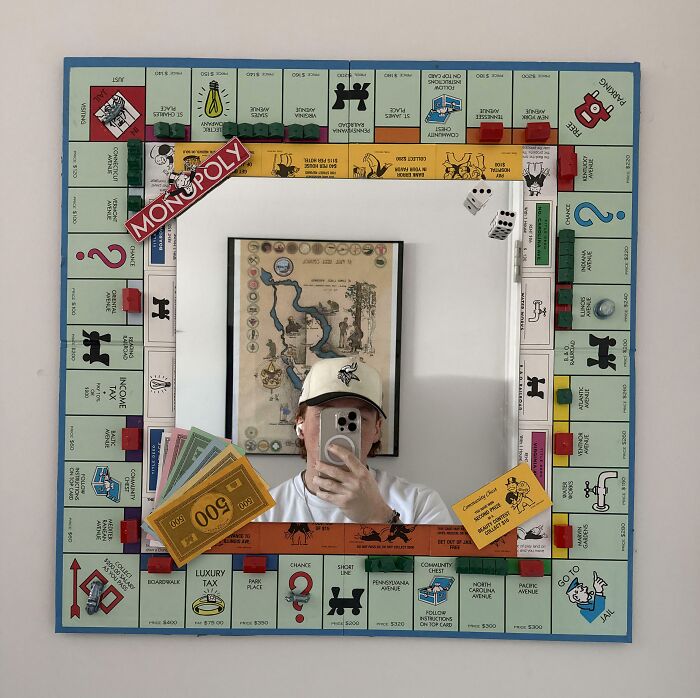 A unique mirror shaped like a Monopoly board, with a person reflected holding a phone, showcasing creative trash into art.