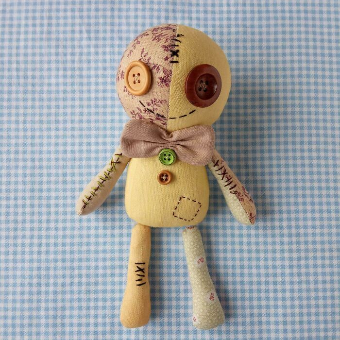 A creative people artistic doll made from trash, featuring a bow tie, button eyes, and mixed fabric patches on a blue gingham background.