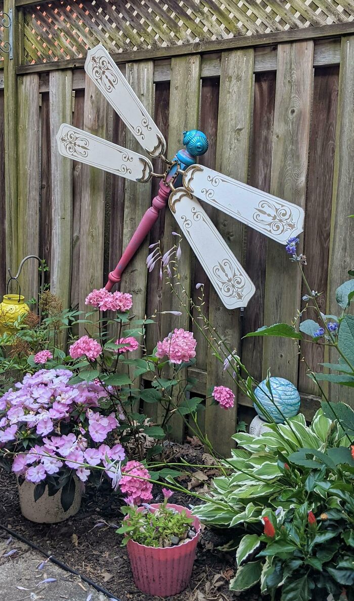 Creative people transforming trash into a dragonfly garden decoration, surrounded by pink flowers against a wooden fence.