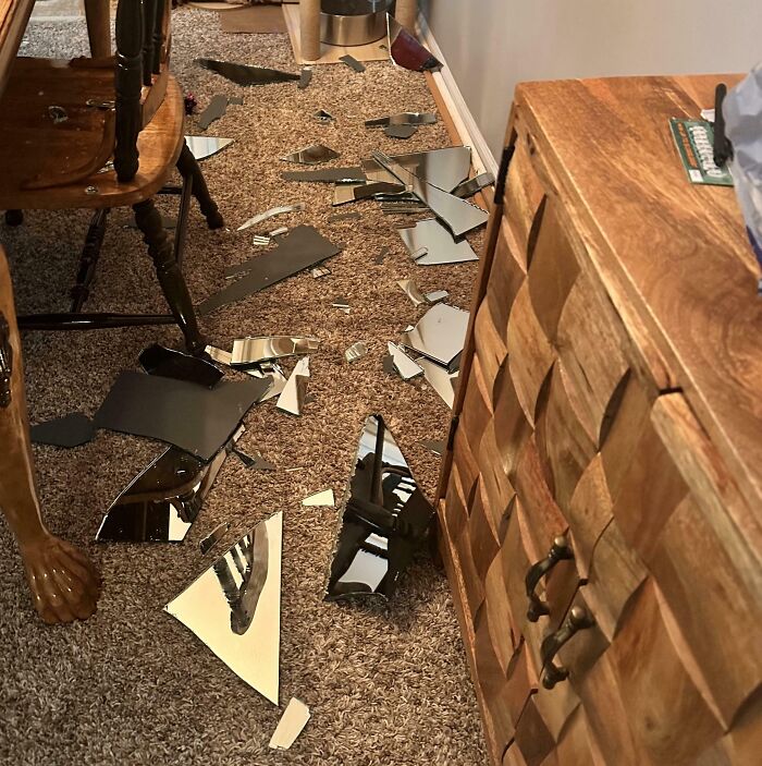 Shattered mirror pieces scattered across a brown carpet, a home improvement fail. Wooden furniture and a chair are also visible.