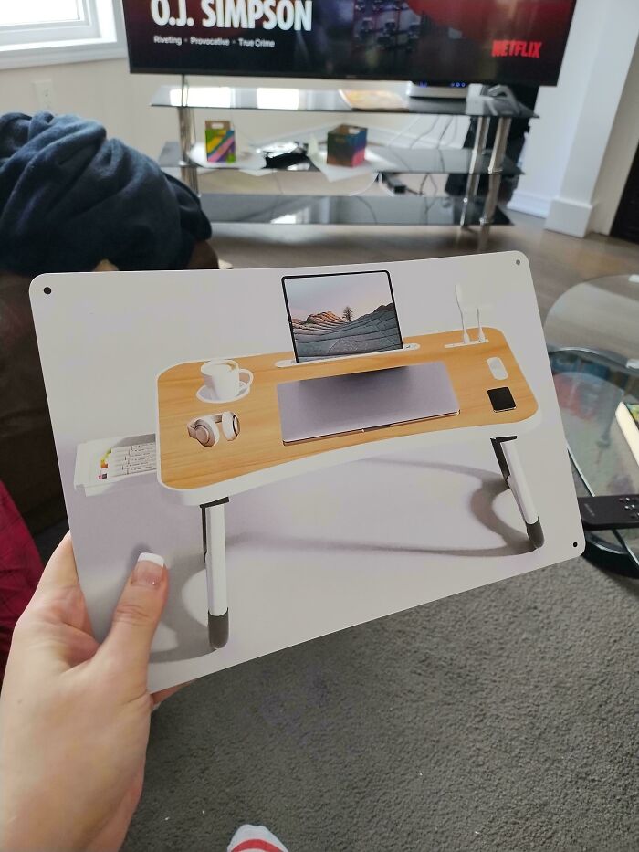 Hand holds a picture of a portable laptop desk with a laptop, tablet, and coffee. Background shows a TV. Temu Or AliExpress purchases.