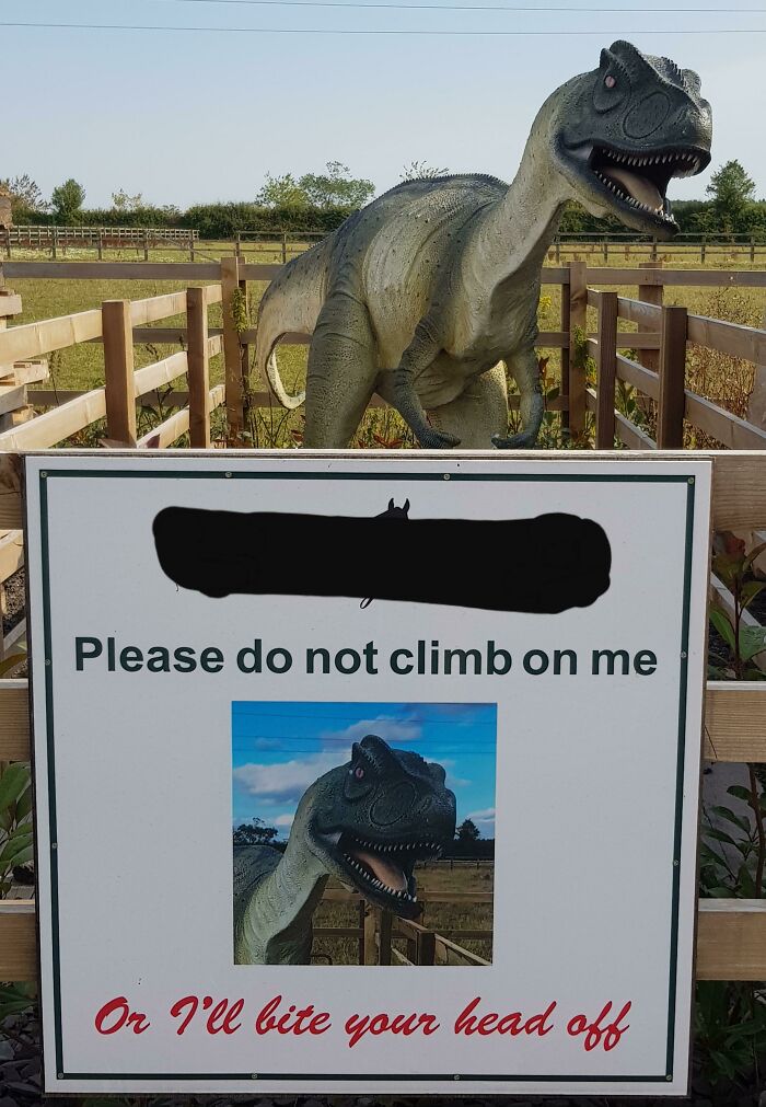 A dinosaur statue in a field with a sign saying, Please do not climb on me, or I'll bite your head off. This makes no sense to city kids.