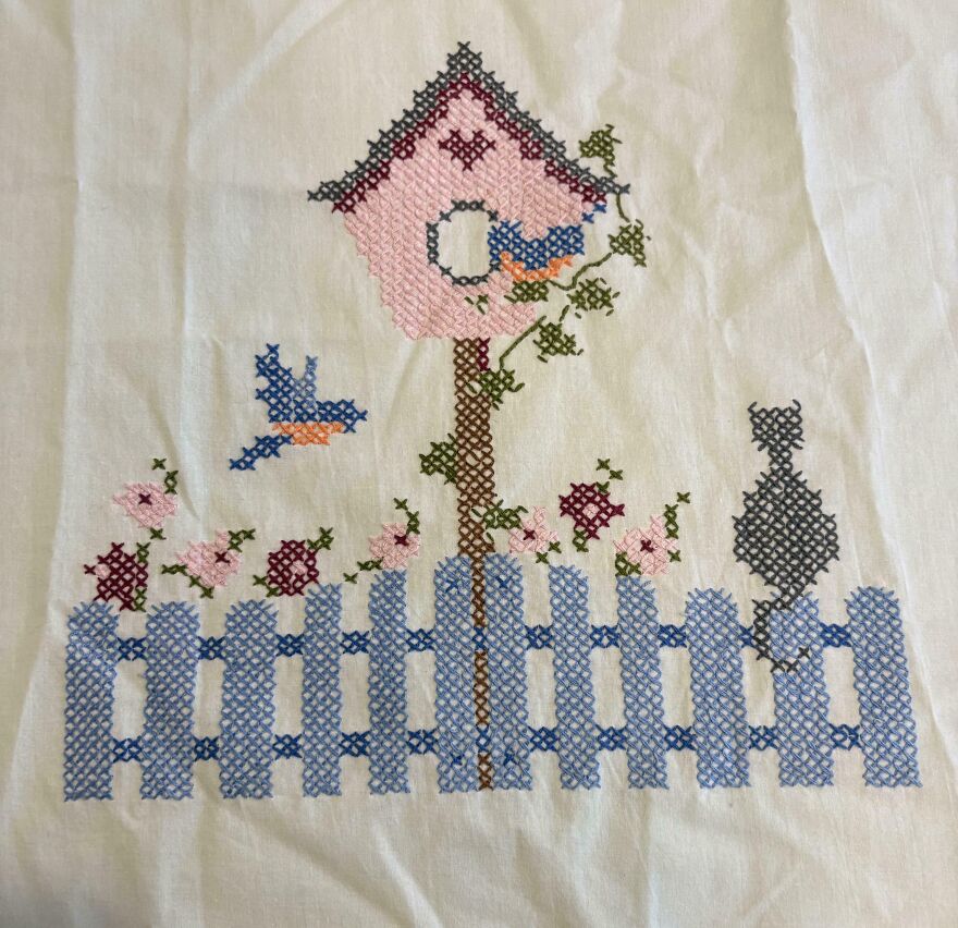 Cross-stitch embroidery on white fabric, showing a pink birdhouse, blue fence, birds, flowers, and a cat. A surprising Goodwill bin find.