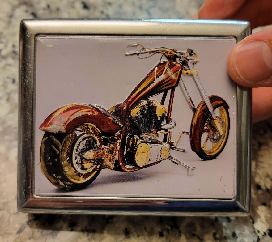 A hand holds a gold bar-shaped lighter case with a custom motorcycle design, a unique Goodwill bin find.