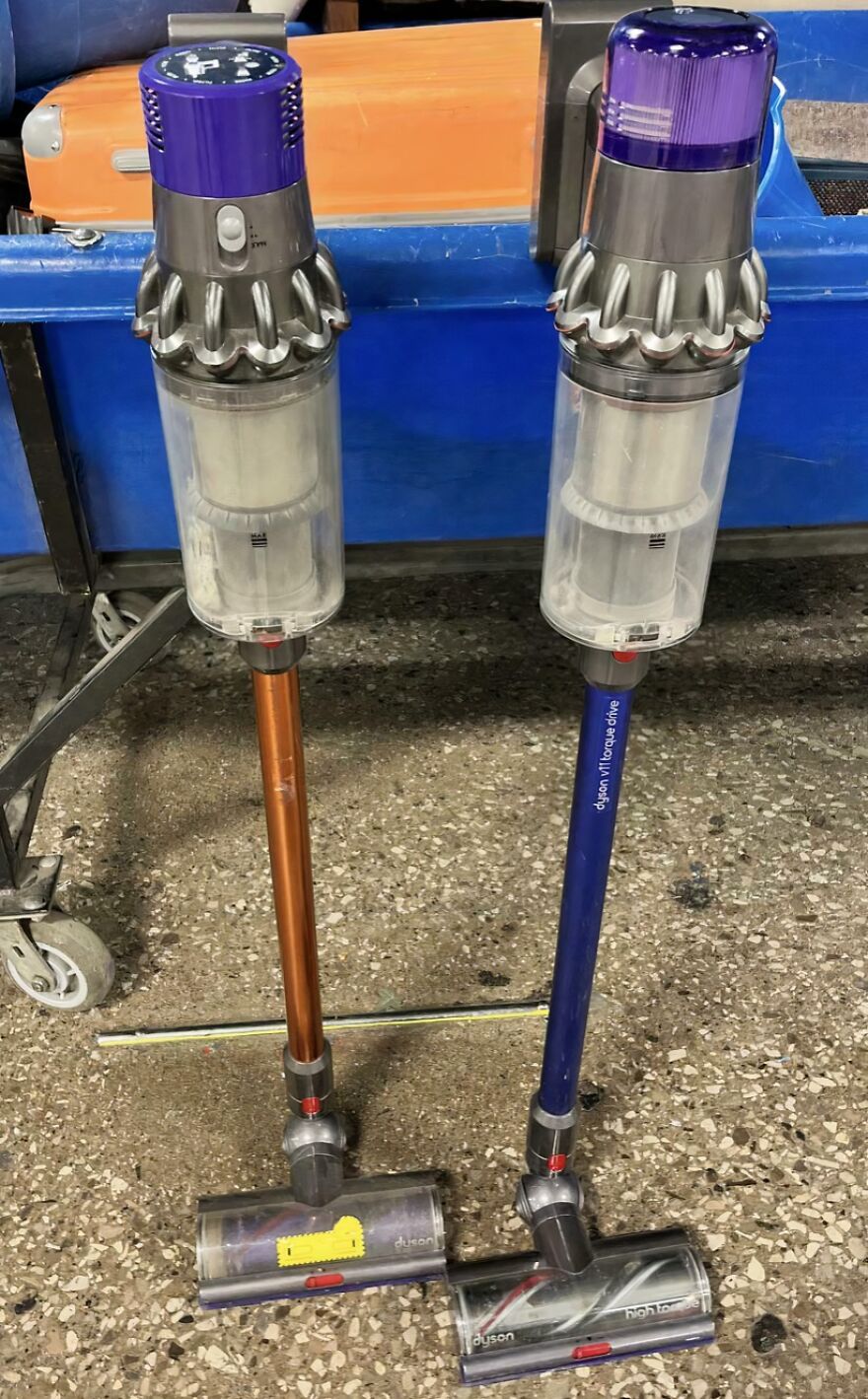 Two Dyson vacuums, one orange and one blue, stand on a concrete floor. A surprising Goodwill bin find.