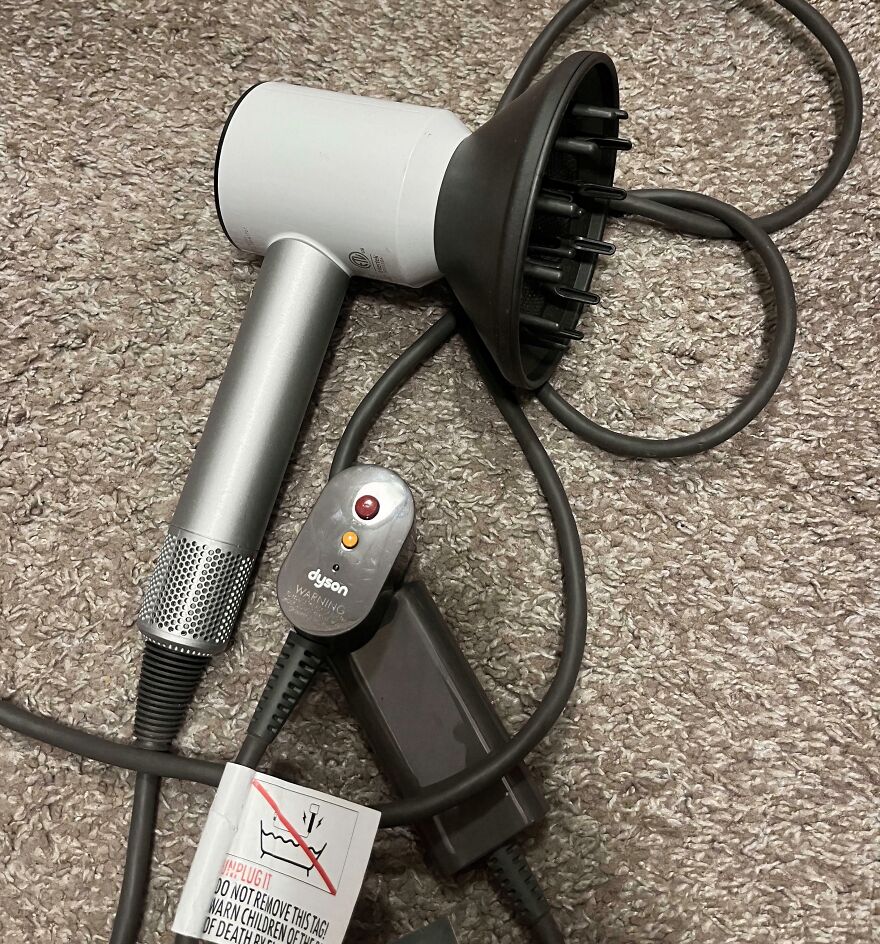 A Dyson hair dryer with a diffuser attachment, cords, and a warning tag on a carpet, a Goodwill find.