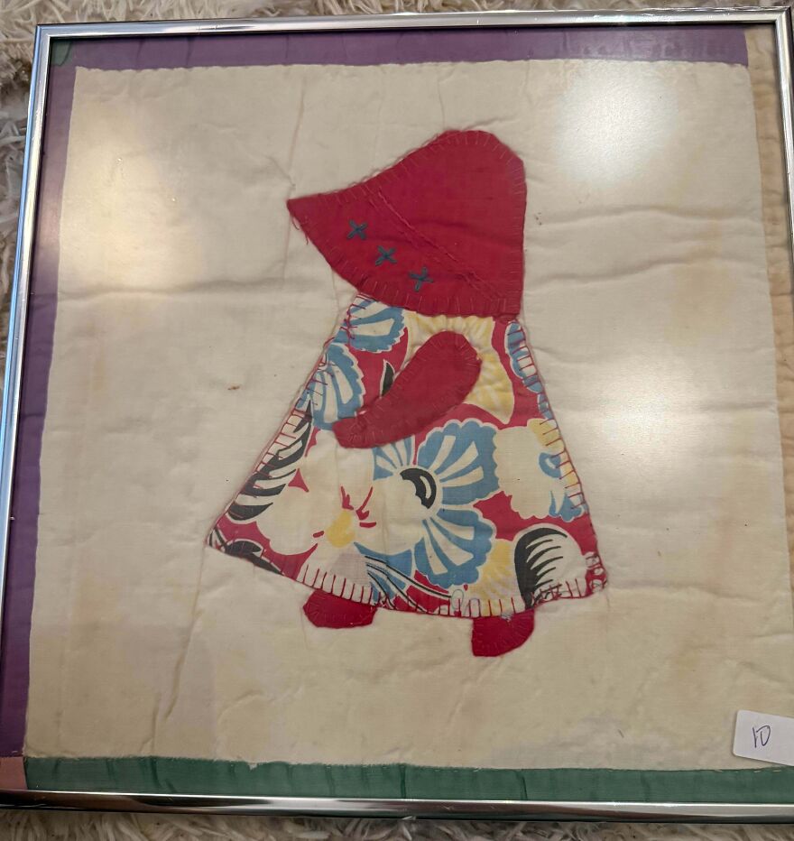 Framed fabric art of a doll in a red bonnet and floral dress, a surprising Goodwill find.