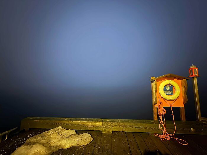 A dark, moody pier extending into vast, uncomfortable water at twilight, with a life preserver station on the right.
