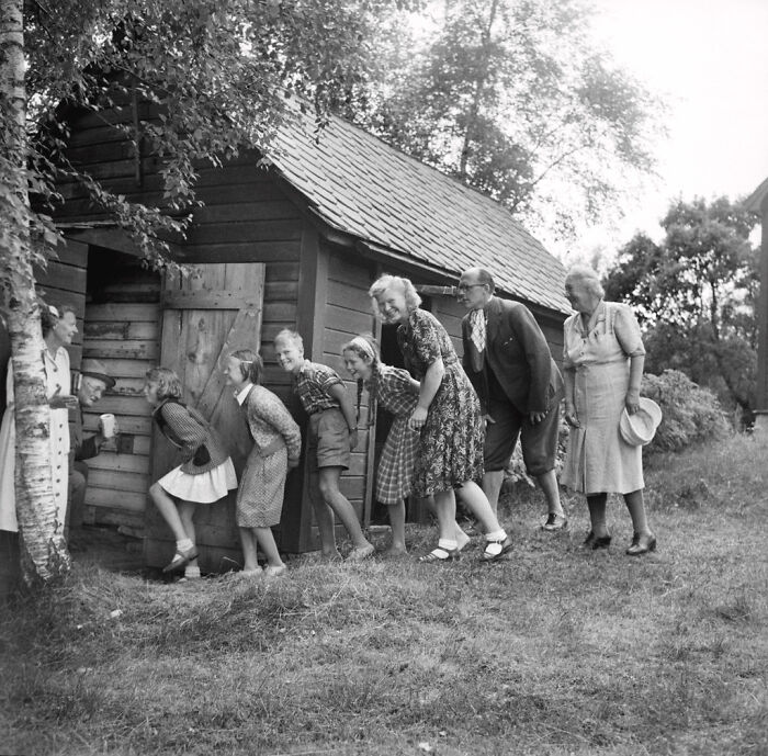 Candid old photo of a large family gathering, everyone is playfully peeking into a small wooden shed.
