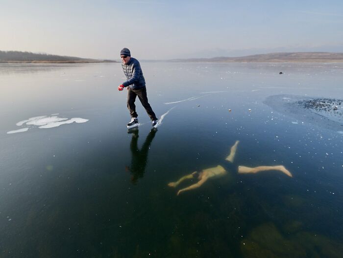 A person iceskating on a frozen lake with another person visible swimming underneath the clear ice. Uncomfortable water pics.