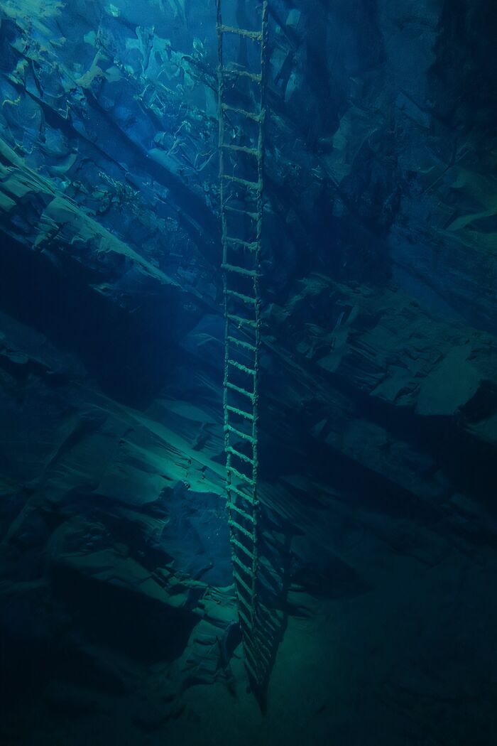 An old ladder descends into deep, dark blue water, surrounded by jagged underwater rock formations, creating an uncomfortable water pic that makes you feel small.