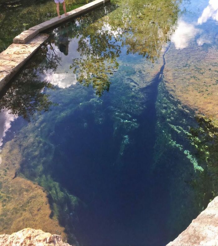 A deep, clear blue natural water pool with visible algae and reflections of trees and clouds, making one feel small.