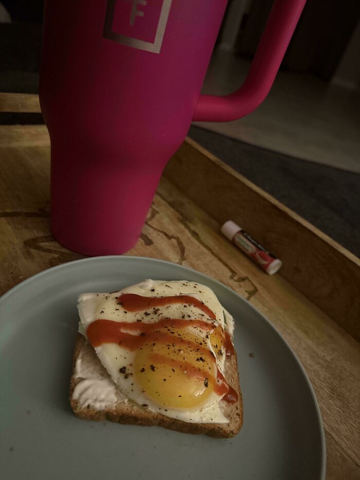 A girl dinner pic: a fried egg on toast with hot sauce and pepper on a plate next to a pink tumbler and lip balm.