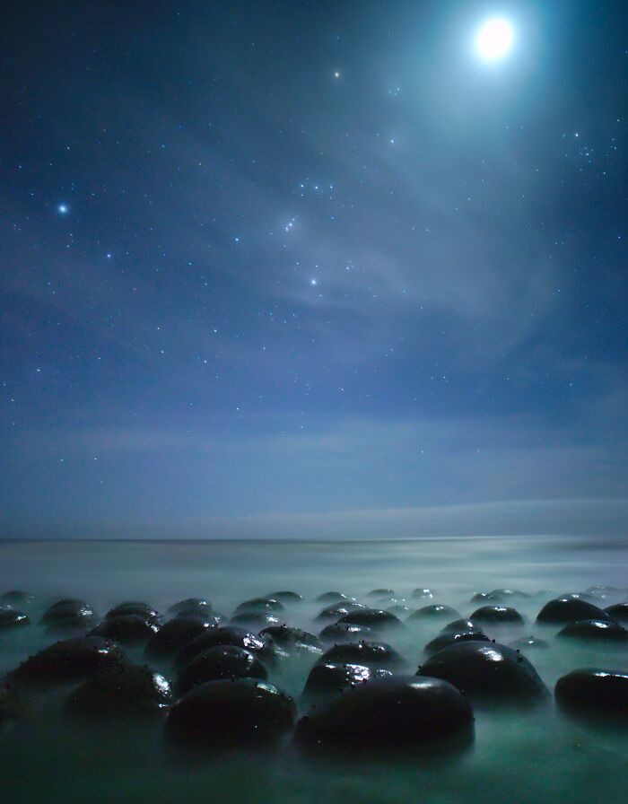 Unbelievable photos taken on Earth: A moonlit night sky over a misty sea with dark, smooth rocks.