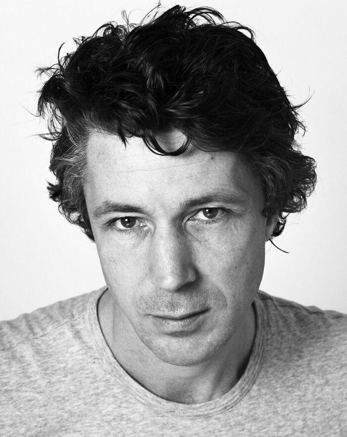 Aidan Gillen, a close-up black and white portrait, looking directly at the camera, showcasing his career highlights.