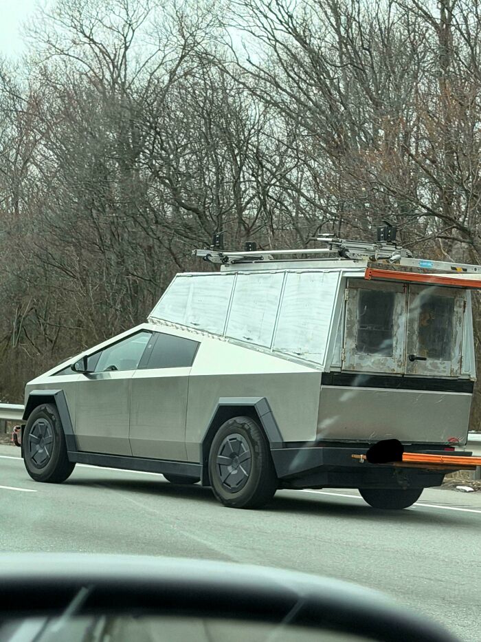 A Tesla Cybertruck with a homemade, DiWHY style rear camper conversion and a ladder rack, on a highway.