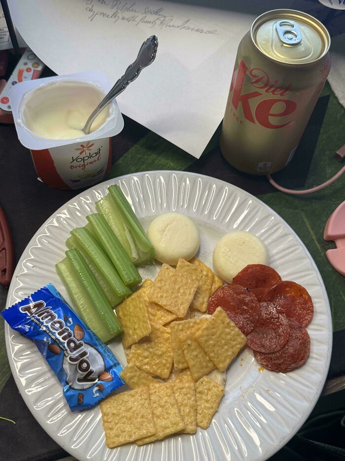 A plate of girl dinner with celery, cheese, crackers, pepperoni, an Almond Joy, yogurt, and a Diet Coke.